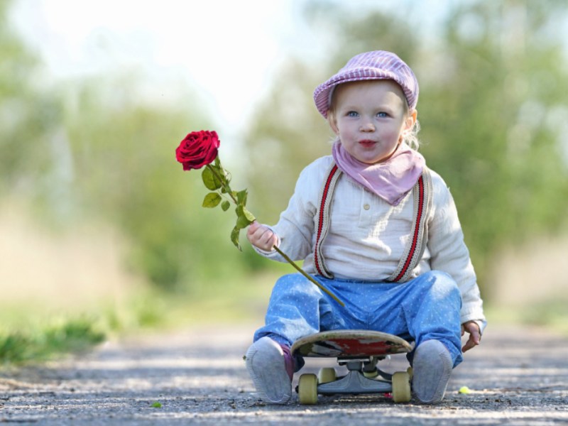 Ein kleiner, schick angezogener Junge mit einer Rose in der Hand sitzt auf einem Skateboard.