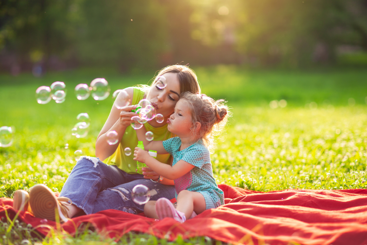 Eine Mutter sitzt mit ihrer Tochter auf einer Wiese und bläst Seifenblasen.