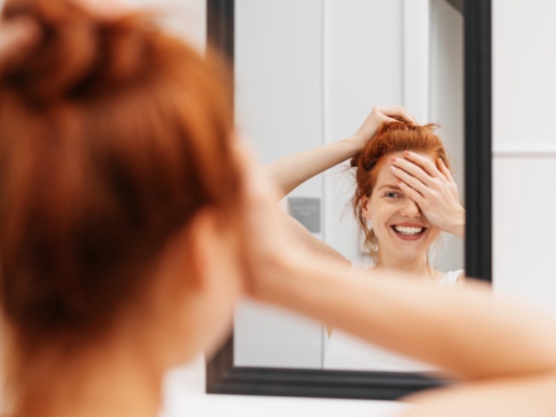 Lachende junge Frau, die sich in einem Spiegel betrachtet, während sie ein Auge mit der Hand abdeckt und sich auf das Spiegelbild konzentriert.