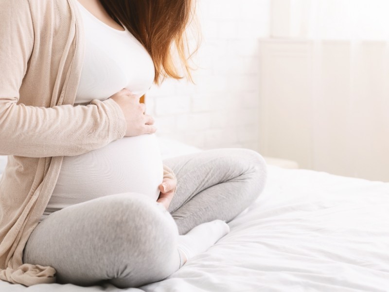 Eine werdende Mutter sitzt auf einem Bett und hält sich den Bauch.