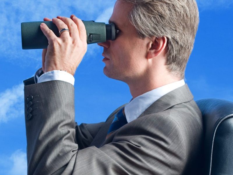 Ein Geschäftsmann schaut mit einem Fernglas in den blauen Himmel.