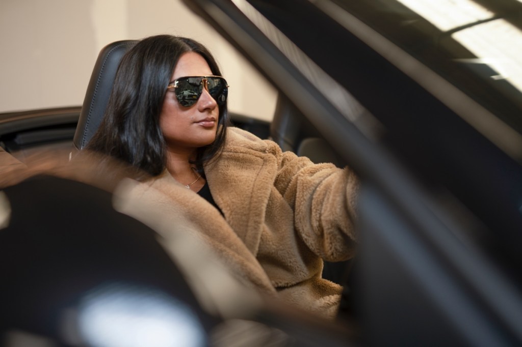 Eine junge Frau mit Sonnenbrille und Pelzmantel sitzt in einem Cabrio.